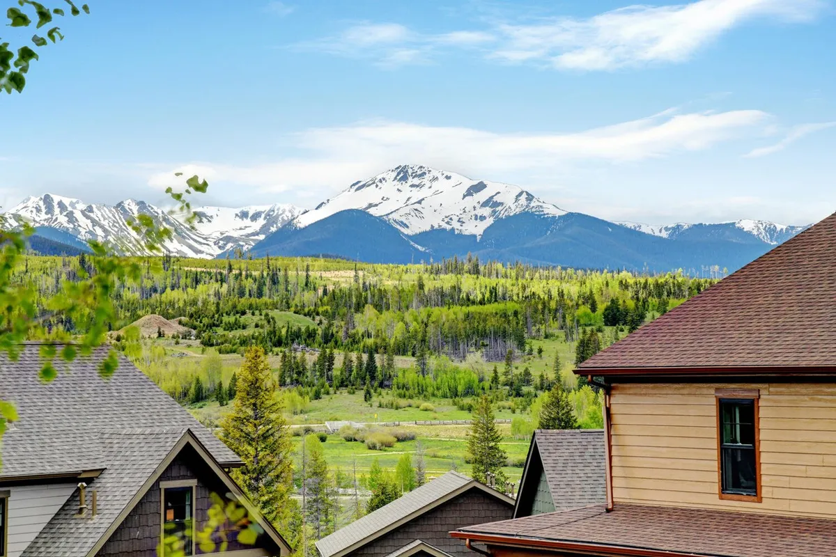 A breathtaking view of snow-capped Rocky Mountain peaks towering above lush green valleys, with rooftops of nearby homes nestled below. The scene exudes the majestic natural beauty and vastness of the surrounding wilderness.