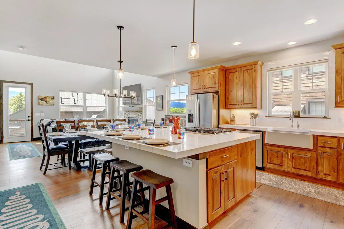 A bright, communal space featuring a generous kitchen island and a large dining table surrounded by high-back chairs. Wooden cabinetry, pendant lighting, and expansive windows fill the space with natural light, creating a cheerful environment ideal for group gatherings and celebrations.