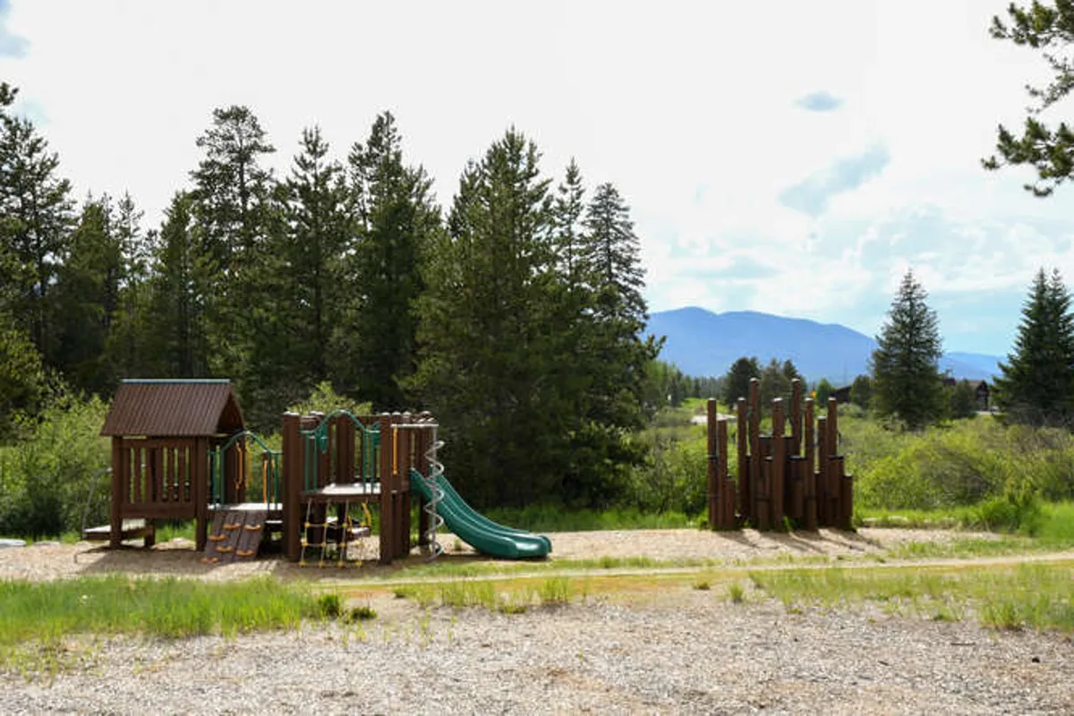 A bright and cheerful community playground with a green slide and tall wooden climbing structure, set against a backdrop of open space. Families and children are shown playing, emphasizing a lively, inviting atmosphere.