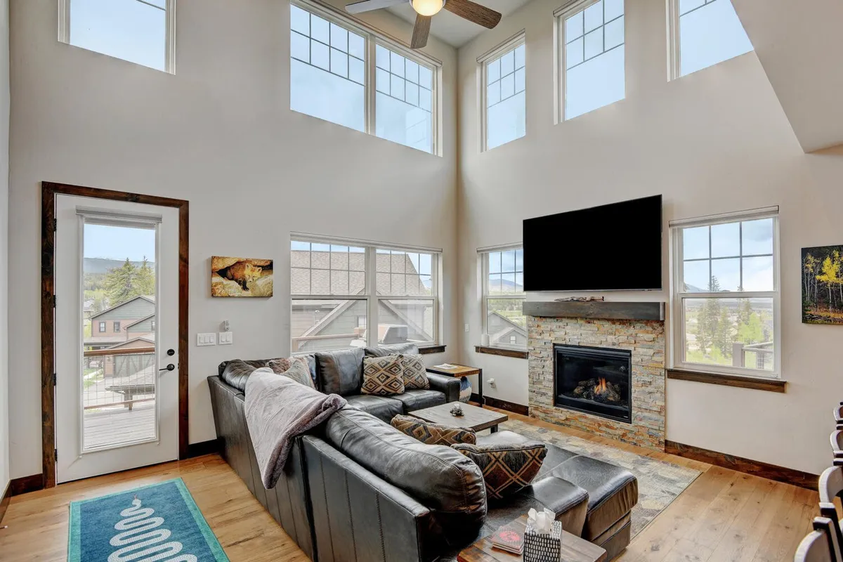 A spacious living room featuring a large sectional sofa facing a stone fireplace, complemented by a large flat-screen television. The room is framed by soaring windows that offer stunning mountain views, flooding the space with natural light.