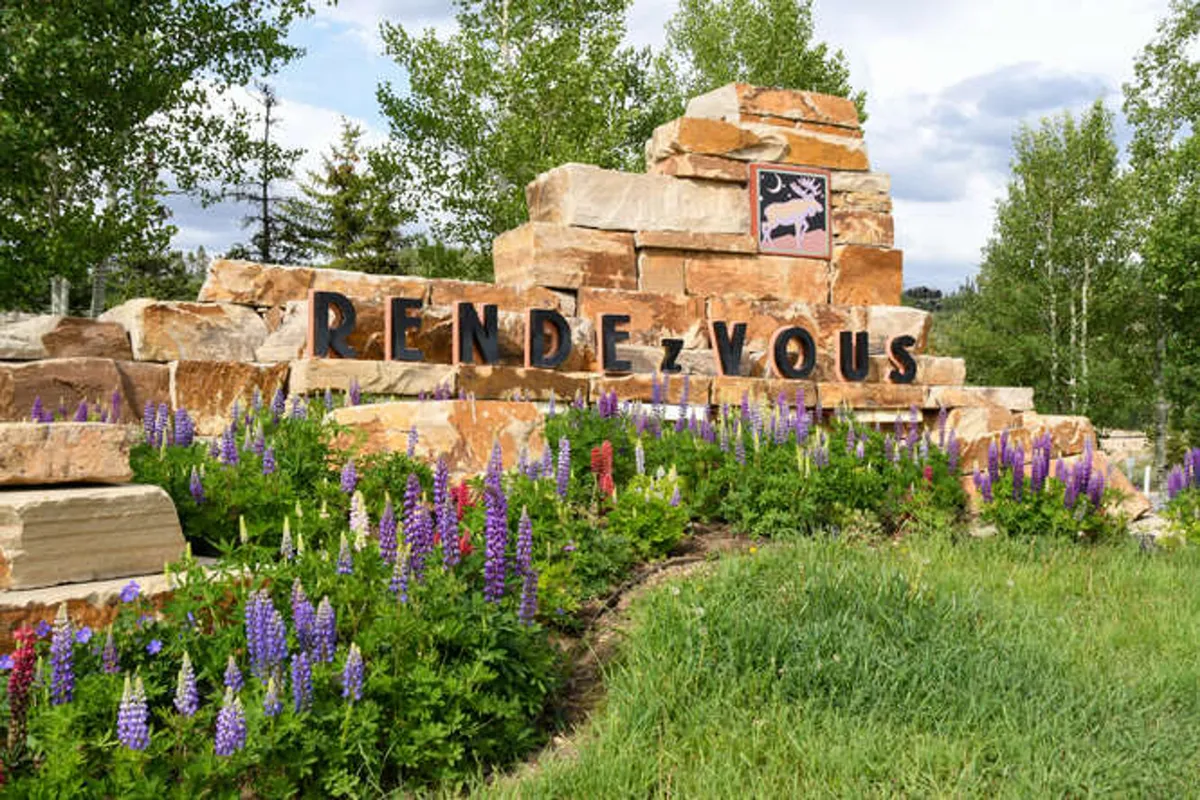 A welcoming stone monument stands amidst vibrant, colorful flowers. The area looks inviting and well-maintained, suggesting a community gathering spot or trailhead, ideal for dog walks and exploration.