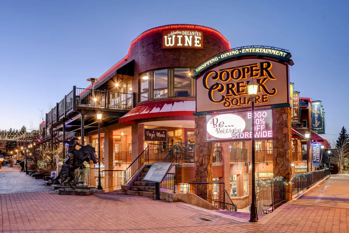 The exterior of Cooper Creek Square features a bold sign, festive lighting, and distinctive architecture. This area is bustling with activity, suggesting a lively downtown with shops, restaurants, and open-air patios.