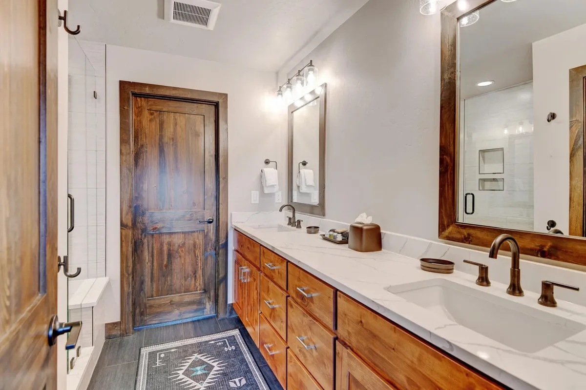 A spacious bathroom with a double vanity, large mirrors, textured rug, and warm wooden cabinetry. The area exudes a calming atmosphere, ideal for self-care routines and relaxation.