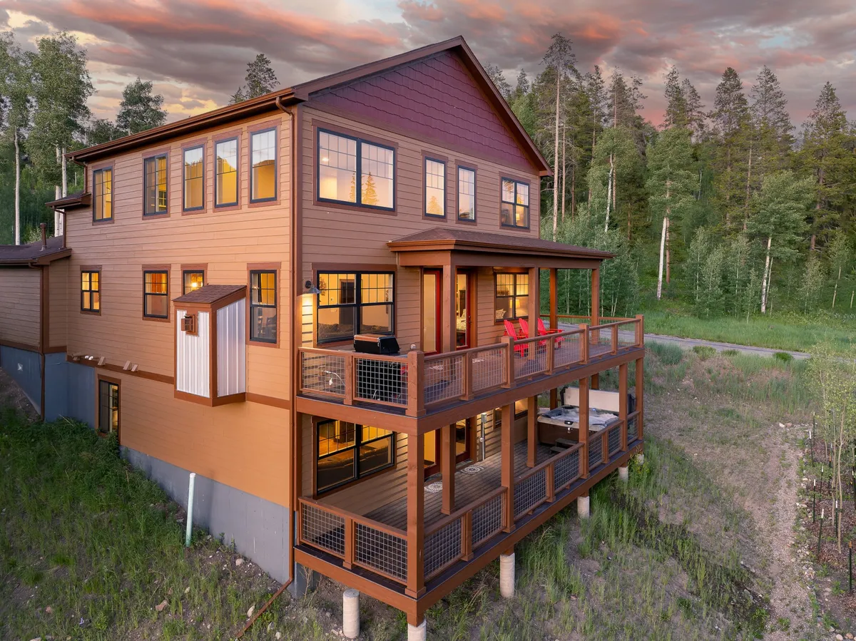 A welcoming deck with comfortable outdoor furniture, large windows connecting to the interior, and lush trees surrounding the space. The scene suggests a seamless flow between indoor and outdoor living, ideal for entertaining or relaxing while enjoying mountain air and views.