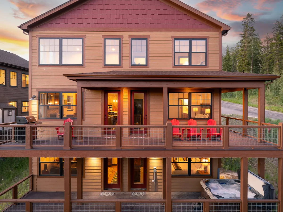 An exterior view of a spacious wooden deck featuring a private hot tub, vibrant red chairs, and large windows that connect the deck with the interior living spaces. The area is surrounded by natural greenery, providing a sense of privacy and relaxation. The hot tub and deck are arranged for both soaking and gathering, perfect for unwinding after outdoor adventures or enjoying summer evenings.