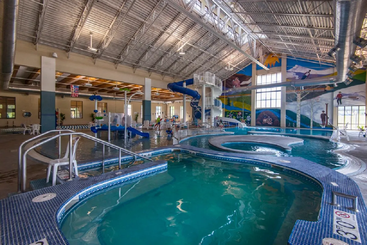 An energetic indoor recreation scene featuring water slides, a swimming pool, colorful murals, and lounge chairs. Children and families are seen enjoying the water attractions, evoking the lively atmosphere of the Grand Park Recreation Center.