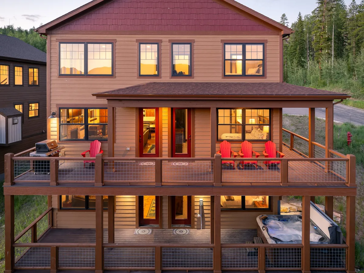 A spacious deck featuring red chairs, a bubbling hot tub, a grill, and expansive windows suggesting the connection between indoor and outdoor living. The warm, welcoming atmosphere is ideal for gatherings, relaxation, and enjoying the mountain setting year-round.