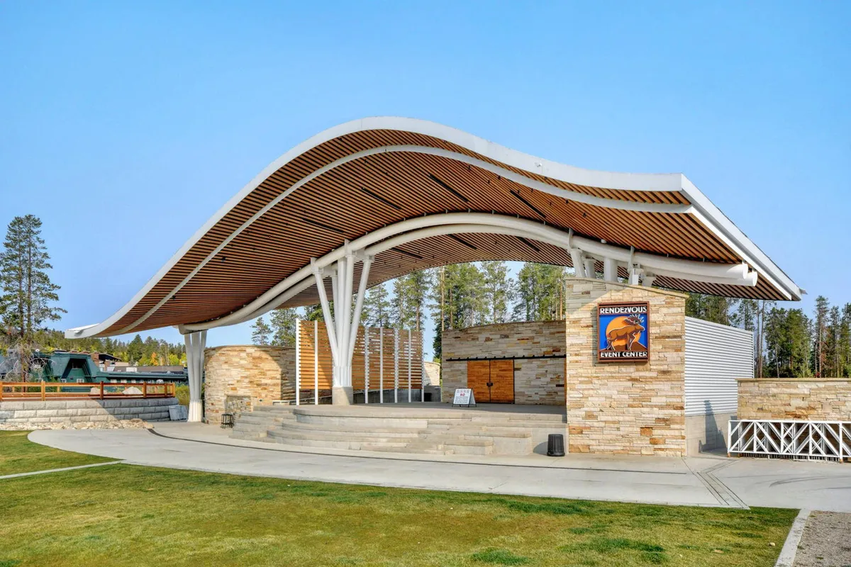 A contemporary outdoor stage and event space with a curved roof structure, stone and wooden architectural accents, and a gathering area perfect for concerts and community festivals. The mood is celebratory and welcoming, ideal for family-friendly gatherings.